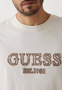 Svijetlosmeđi majica s okruglim izrezom, s tamnosmeđim izvezenim tekstom "GUESS EST.1981". Tkanina djeluje mekano i ima glatku teksturu.