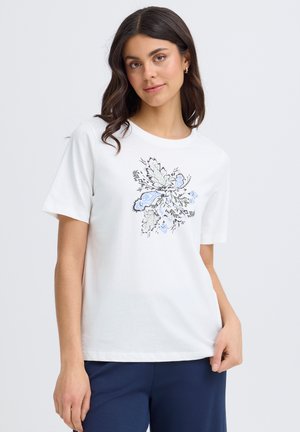 Femeie cu păr lung și închis la culoare, purtând un tricou alb cu un design floral albastru și gri, stând în fața unui fundal gri deschis.