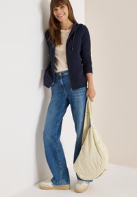 Smilende kvinde iført navy hoodie, beige top, blå jeans og sneakers, bærende en stor beige skuldertaske, stående op mod en tofarvet væg.