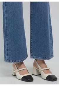 Des chaussures slingback couleur crème avec un bout noir, un talon block et une bride à la cheville, accompagnées d'un jean bleu à jambes larges.