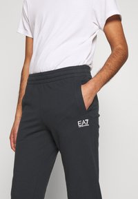 Tmavě šedé jogger kalhoty vyrobené z měkkého materiálu, s elastickým pasem, bočními kapsami a bílým logem na levém bokě.