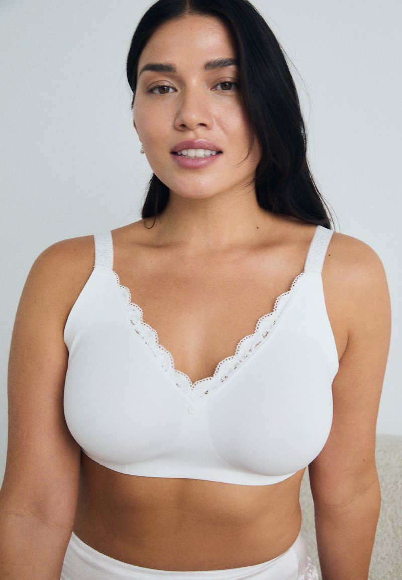 Femme portant un soutien-gorge à bonnets mous blancs avec des détails en dentelle, regardant vers l'avant sur un fond clair et uni.