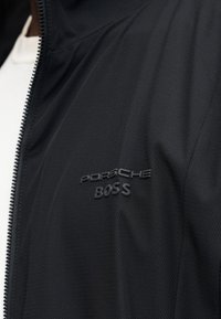 Μαύρο μπουφάν με φερμουάρ, με ανάγλυφο μαύρο λογότυπο "PORSCHE BOSS" στο στήθος, φορεμένο πάνω από άσπρο πουκάμισο.