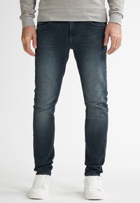 Hombre que lleva vaqueros oscuros de corte slim desgastados, sudadera gris clara y zapatillas blancas, de pie contra un fondo blanco liso.