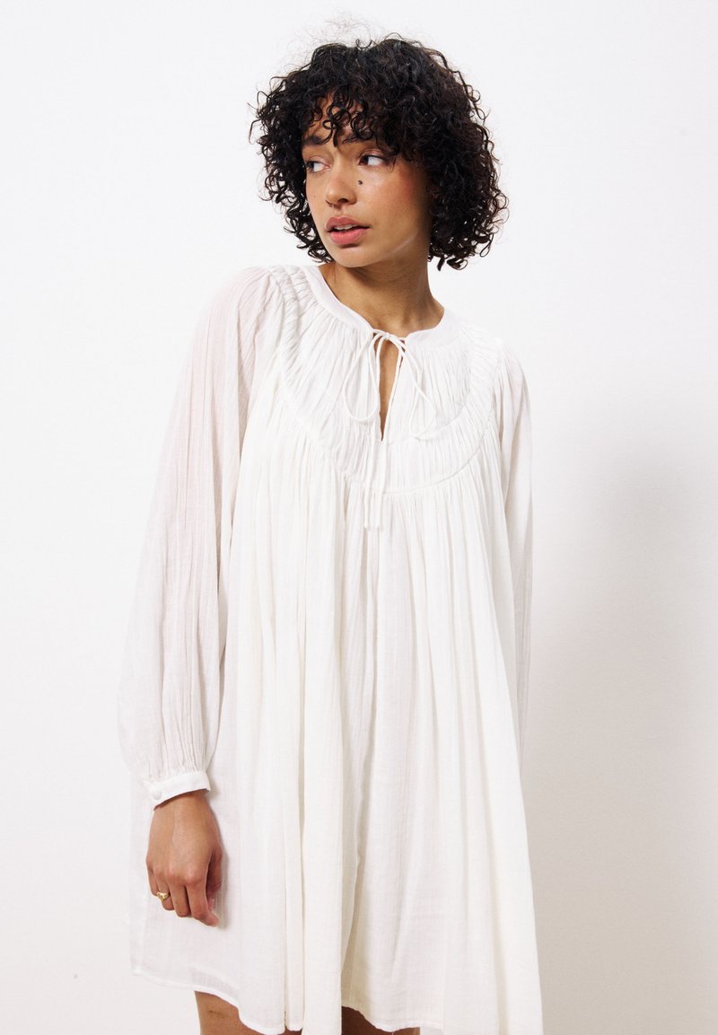 Weißes, locker sitzendes Kleid mit langen Ärmeln, gerafftem Ausschnitt und Bindeband. Leichter Stoff mit einem weichen, strukturierten Erscheinungsbild.