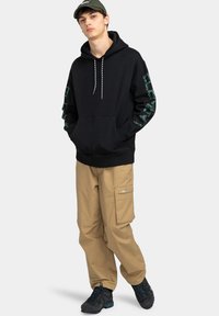 Sudadera negra con diseños gráficos en las mangas, cordón, y bolsillo frontal. Combinada con pantalones cargo beige que tienen bolsillos laterales y tejido texturizado.