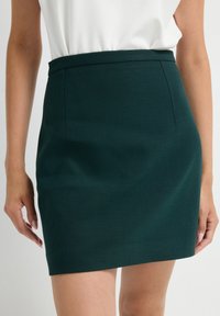 Gonna a matita verde con una texture liscia, caratterizzata da una vita aderente e un design minimale, che arriva a metà coscia. Linee pulite e senza decorazioni.