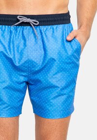 Pantaloni da nuoto blu con pois bianchi, dotati di una cintura elastica e dettagli con cordino. Il materiale sembra leggero e liscio.