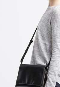 Zwarte leren schoudertas met een klep en rits, voorzien van een gladde textuur, verstelbare band en hardware in goudkleur.