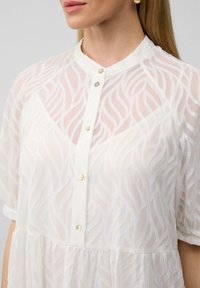 Blusa blanca con patrón de olas texturizadas, mangas cortas y frente abotonado. Hecha de un material ligero y transparente.