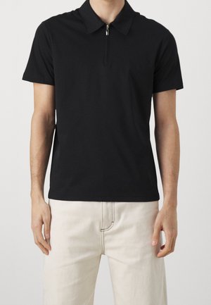 Persona con una camiseta polo negra de manga corta con cremallera frontal y pantalones beige claro sobre un fondo sencillo.