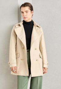 Beige dubbelzijdig jasje met een kraag en schoudernaleidingen, voorzien van goudkleurige knopen en ritszakken. Zachte stoffen textuur.