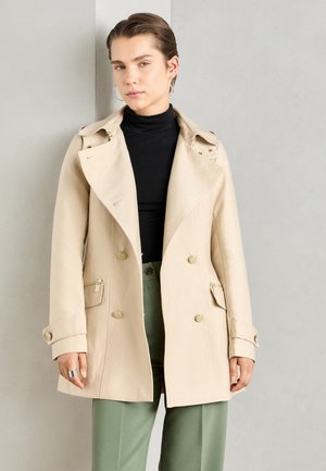 Manteau beige à double boutonnage avec col et épaulettes, doté de boutons dorés et de poches zippées. Texture de tissu douce.