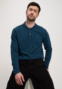 Długo rękawowy polo w ciemnym odcieniu teal z bawełny, z kołnierzykiem, zapięciem na trzy guziki oraz subtelnym logo na klatce piersiowej. Czarne spodnie.