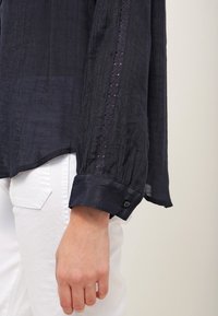 Main portant une chemise à manches longues sombre avec un détail en dentelle et un poignet boutonné, reposant à côté d'un pantalon blanc avec une poche arrière visible.