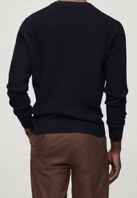 Námořnický žebrovaný svetr s dlouhými rukávy a kulatým výstřihem, s texturovanou tkaninou a těsným střihem v oblasti spodního lemu, spárovaný s hnědými kalhotami.