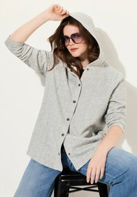 Svetle šedá mikina s kapucí, s texturovaným vzorem, snap knoflíky a dlouhými rukávy. V kombinaci s modrými džíny a oversized slunečními brýlemi.