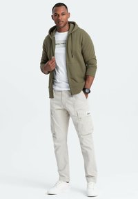 Olivgrüner Zip-Hoodie über einem weißen T-Shirt, kombiniert mit hellbeigen Cargo-Hosen und weißen Sneakern. Verfügt über Fronttaschen und einen lässigen Schnitt.