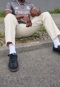 Grå sportströja med vita accenter, krämfärgade byxor och svarta sneakers. Vita strumpor synliga med ett rött accentband. Sittande position.