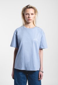 Camiseta de algodón azul claro con cuello redondo y mangas cortas. Presenta un logo bordado blanco en el lado izquierdo del pecho. Llevada con pantalones de mezclilla de cintura alta.