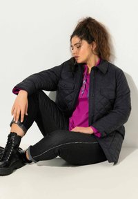 Mujer sentada en el suelo con una chaqueta de quilting negra, sudadera morada, jeans negros remangados y botas negras con cordones, contra una pared blanca.