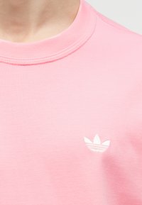 Κοντινό πλάνο ενός ατόμου που φοράει μια ανοιχτό ροζ μπλούζα με στρογγυλή λαιμόκοψη και μικρό λευκό λογότυπο Adidas trefoil στο στήθος.