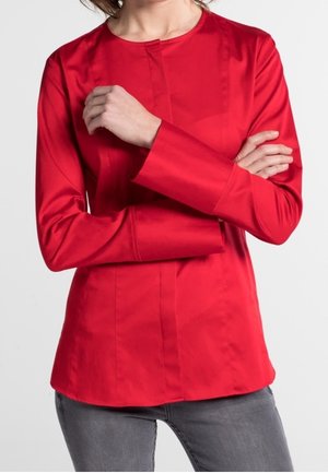 Kobieta w jasnoczerwonej bluzce z długim rękawem, okrągłym dekoltem i ukrytymi guzikami z przodu, w połączeniu z szarymi dżinsami.