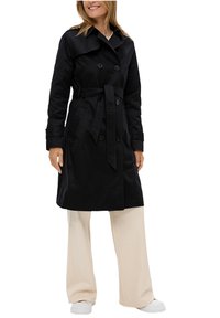 Manteau noir avec boutons double-breasted, taille ceinturée et large col, associé à un pantalon large de couleur crème.