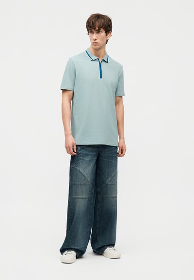 Jeune homme portant un polo à manches courtes bleu clair avec un col rayé, un jean large en denim foncé et des baskets blanches, debout et regardant vers la gauche.