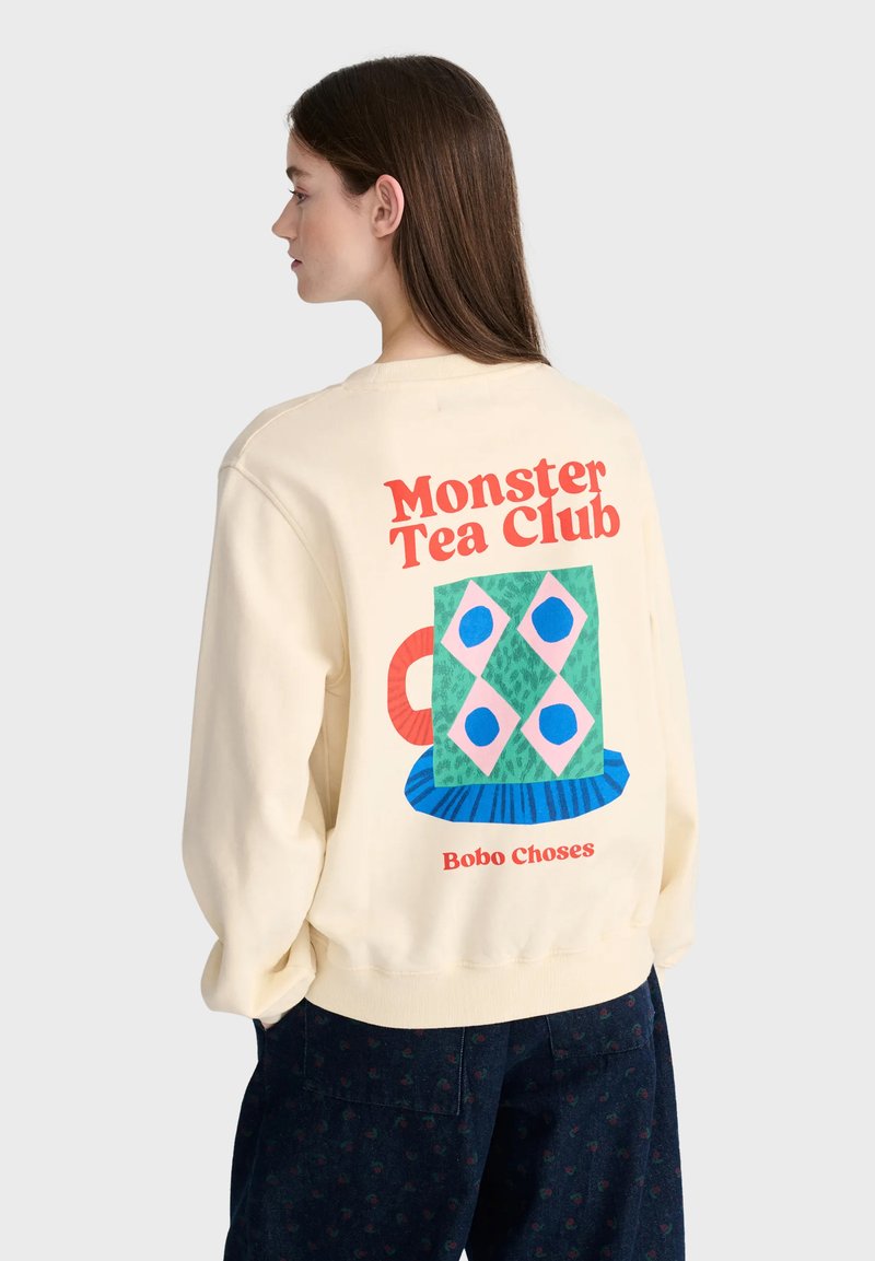 Jeune femme aux longs cheveux portant un sweat-shirt couleur crème avec le texte "Monster Tea Club" et un motif coloré de tasse géométrique dans le dos.