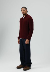 Man met gevlochten haar, gekleed in een bordeauxrode gebreide trui, donkerblauwe spijkerbroek en bruine wandelschoenen, staand tegen een effen lichte achtergrond.