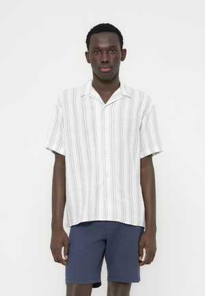 Κοντομάνικο πουκάμισο σε ανοιχτό μπεζ με κάθετες ρίγες, κατασκευασμένο από ελαφρύ ύφασμα, συνδυασμένο με σορτς σε ναυτικό μπλε χρώμα.
