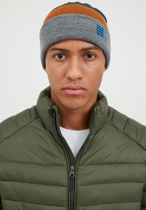 Knitted beanie in gray, featuring orange and navy stripes. Worn with a green puffer jacket, highlighting a zip closure and quilted texture.