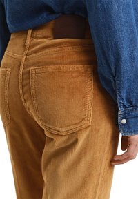 Bruine corduroy broek met een ribbeltextuur; heeft twee achterzakken en een leren merklabel op de taille. Blauwe spijkerblouse in de buurt.