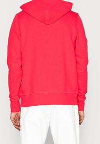 Red hooded sweatshirt with a soft texture, featuring a kangaroo pocket and ribbed cuffs. The back has a clean, minimal design.