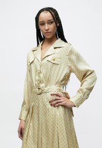 Μπεζ πουκαμισοφόρεμα με διακριτικό επαναλαμβανόμενο μοτίβο, εμπρόσθια με κουμπιά, ραφτό γιακά, πλαϊνές τσέπες και ζώνη στη μέση που ταιριάζει.