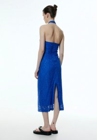 Blaues Midikleid aus Spitze mit Halterneck, offenem Rücken und seitlichem Schlitz. Der Stoff hat eine strukturierte Oberfläche mit floralen Mustern und ist figurbetont.