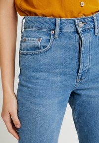 Korkeavyötäröiset siniset farkut, joissa on sileä rakenne, kupariset niitit ja perinteinen viiden taskun muotoilu.