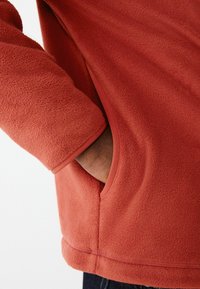 Fleece-Pullover in verbranntem Orange mit weicher Textur. Verfügt über eine Seitentasche und kontrastierende Besatz entlang der Kanten und Bündchen.