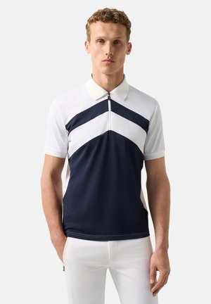 Man wearing short-sleeve white and navy blue zip-up polo shirt with chevron pattern, paired with white pants, standing against plain background.