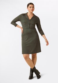 Vestido de patrones en negro y verde azulado con cuello tipo camisa, mangas de tres cuartos y silueta ajustada, combinado con botas negras hasta el tobillo.