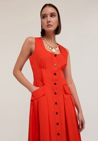 Rochie din bumbac roșu, cu un design fără mâneci, decolteu pătrat, butoane pe față, buzunare laterale și detalii pliate în poală.