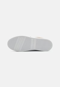 Biała podeszwa sneakerów z wzorowanymi bruzdami, z logo w kolorze złotym. Gładka faktura, zaokrąglony kształt i solidny materiał gumowy.