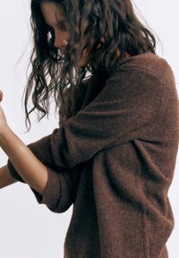 Žena s dugom kovrčavom kosom koja nosi smeđi džemper, okrenuta bočno s savijenim rukama, na jednostavnoj svijetloj pozadini.