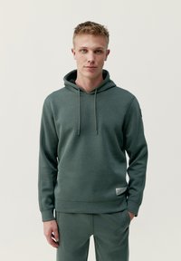 Zelená mikina s kapucňou vyrobená z mäkkého materiálu, s nastaviteľnou kapucňou, rebrovanými manžetami, prednou vreckom a malou etiketou na leme.