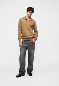 Καφέ φούτερ με φερμουάρ και ριμπ μανσέτες, σε συνδυασμό με χαλαρά εφαρμοστά γκρι τζιν και μαύρα παπούτσια. Το μοντέλο στέκεται μπροστά σε λευκό φόντο.