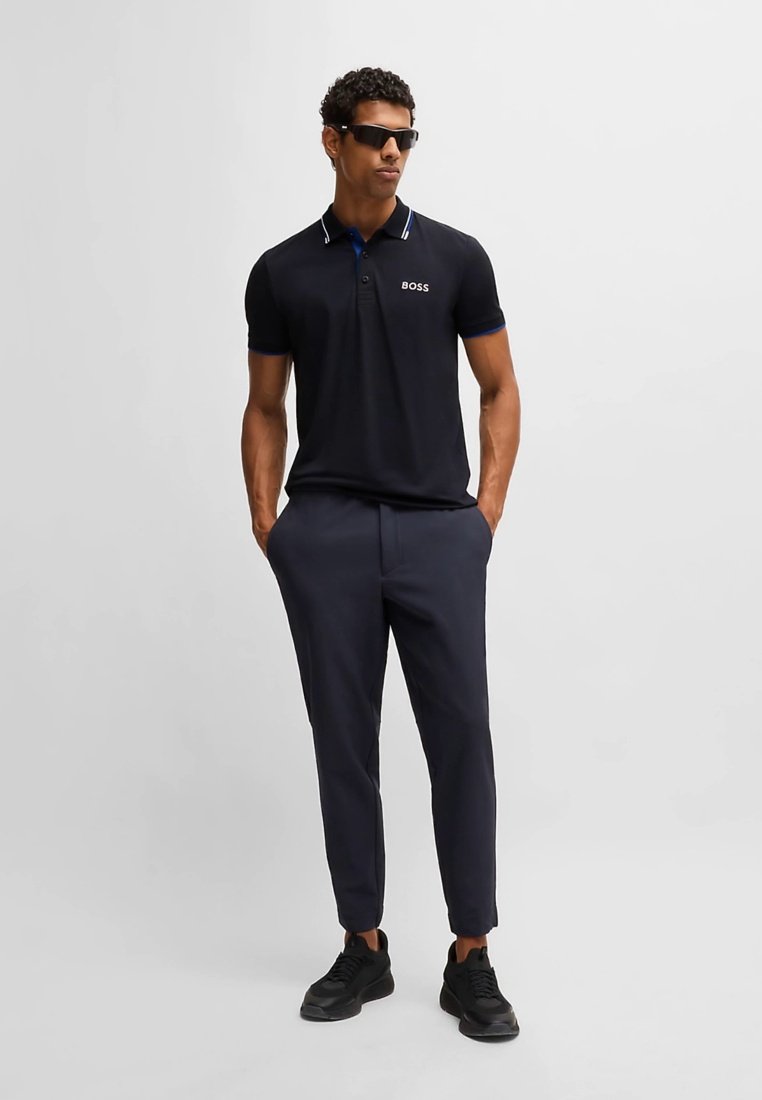Polo noir avec accents bleus et logo blanc, associé à un pantalon foncé et des baskets noires. Des lunettes de soleil ajustables complètent le look.