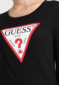 Zwarte lange mouw shirt met een driehoekig rood en wit logo met "GUESS," een vraagteken en numerieke details op een gestructureerde stof.
