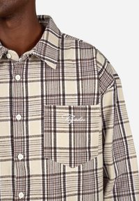 Brun og kremruter skjorte med krage som kan kneppes ned, frontlomme og hvit logo-detalj på lommen. Laget av stoff med teksturert overflate.