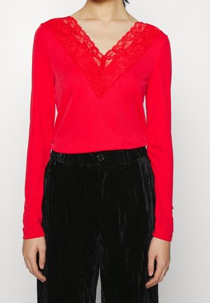 Femme portant un haut rouge vif à manches longues avec un détail en V en dentelle et un pantalon en velours noir, debout devant un fond blanc.